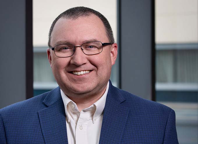 A man wearing glasses, a blue blazer, and a white shirt smiles at the camera in front of large windows.