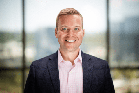 A smiling man in a business suit stands indoors in front of large windows with an out-of-focus cityscape in the background.