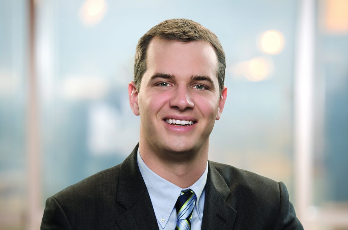A man in a dark suit and striped tie is smiling at the camera in an office setting with a blurred background.