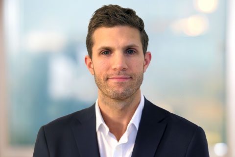 Dark haired man in a black blazer smiles in front of a soft blurred background