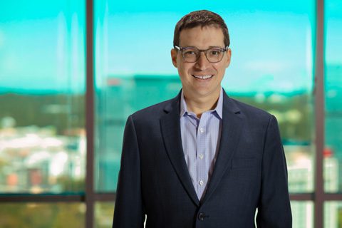 A man wearing glasses and a dark suit jacket over a light blue shirt stands indoors in front of large windows with an urban view in the background.
