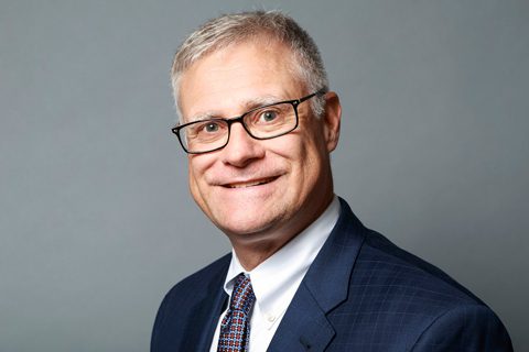 A middle-aged man with short gray hair, glasses, and a suit, smiling at the camera against a plain gray background.