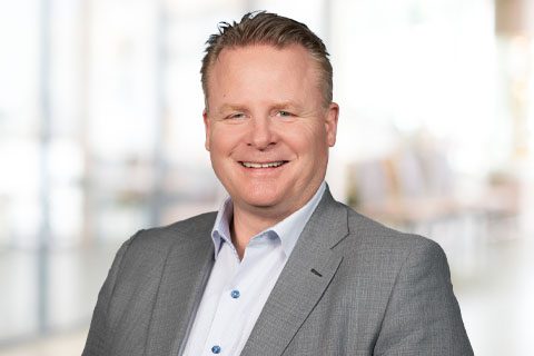 A smiling man in a light gray suit jacket and white shirt stands in front of a blurred indoor office background.