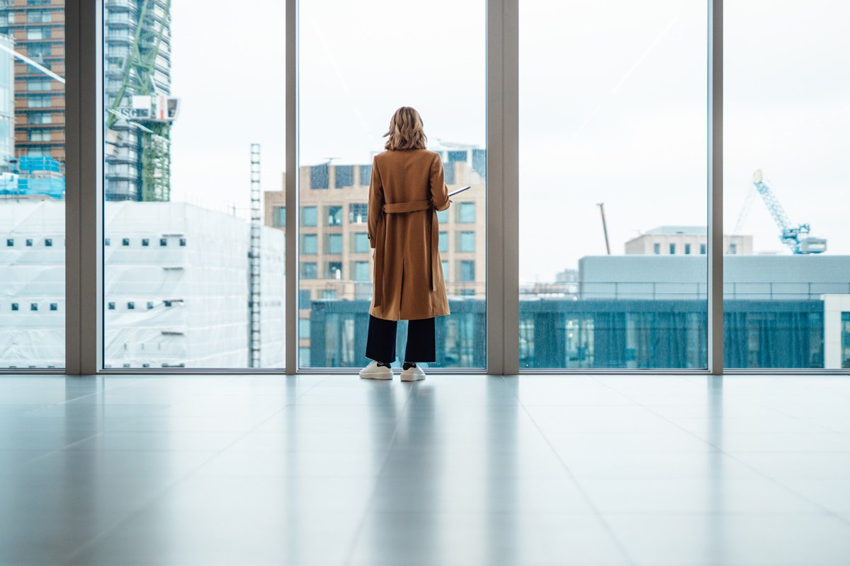 A person wearing a brown coat stands in front of large floor-to-ceiling windows, looking out at city buildings and construction cranes.