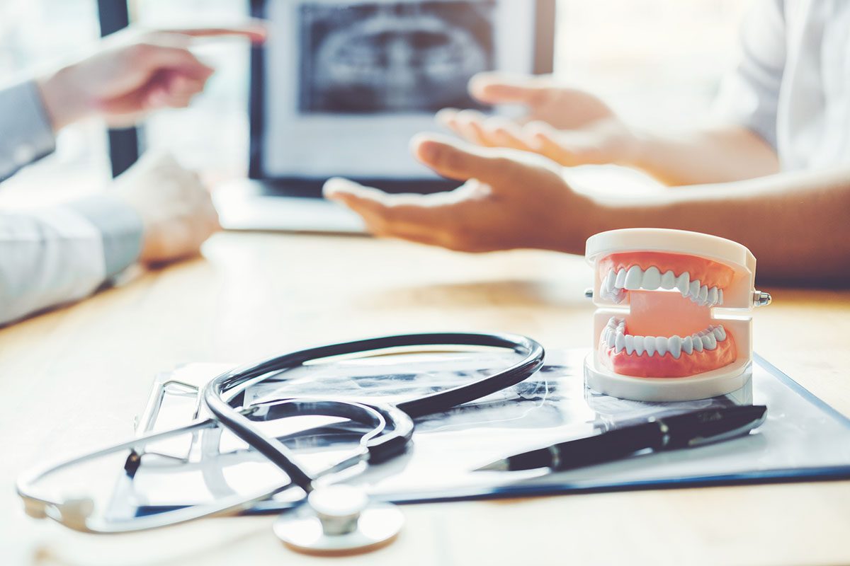 Two people discuss a dental X-ray on a screen; a stethoscope, dental model, and pen are on the table in the foreground.