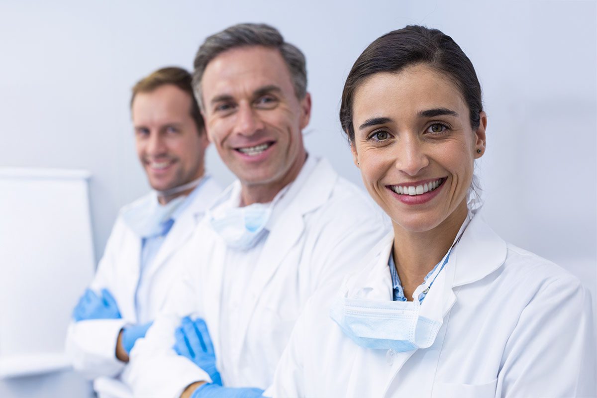 Three smiling medical professionals wearing white lab coats and face masks stand in a clinical setting, looking towards the camera.
