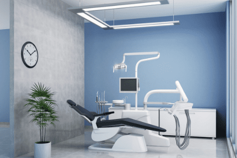 Modern dental office with a reclining dental chair, equipment, a computer monitor, blue walls, a clock, a plant, and bright overhead lighting.