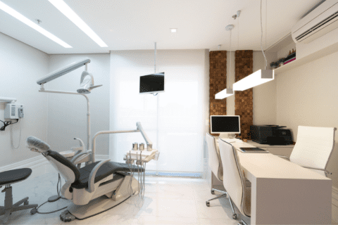 A modern dental office with a dental chair, computer desk, overhead light, wall-mounted monitor, and white furnishings.