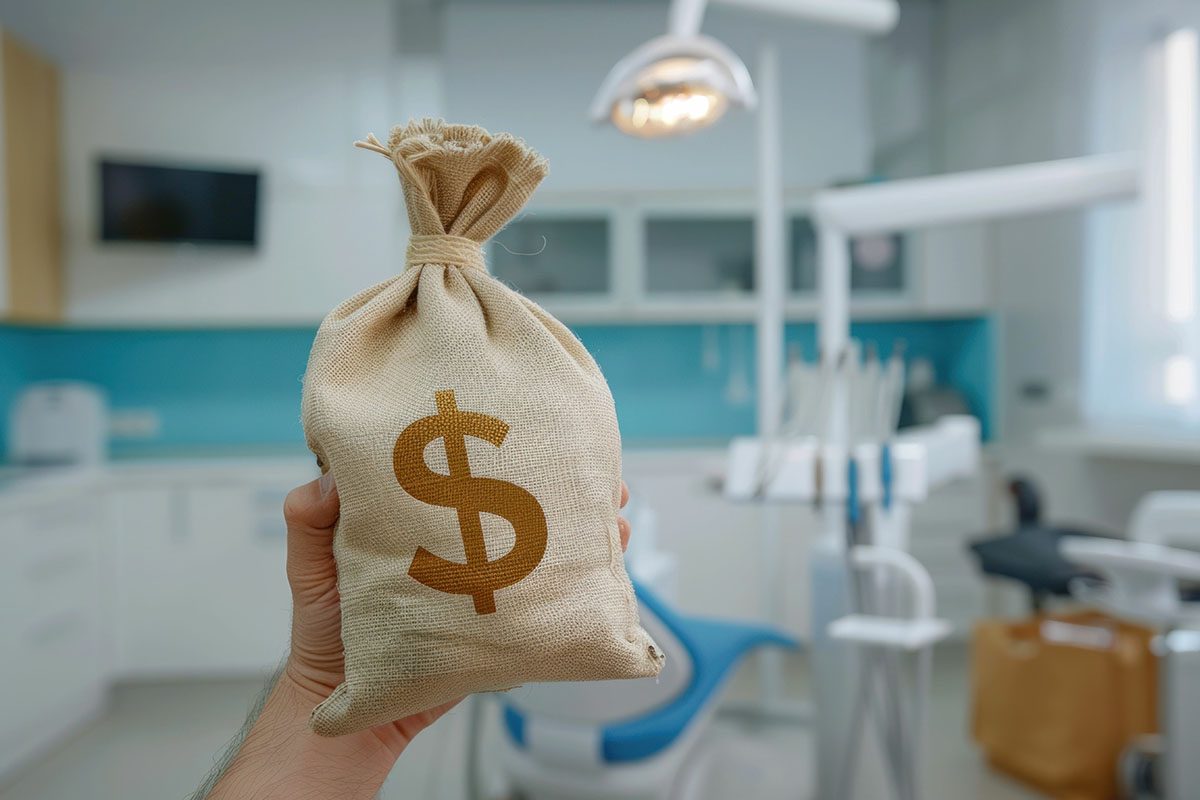 A hand holds a burlap money bag with a dollar sign in a modern dental clinic examination room.
