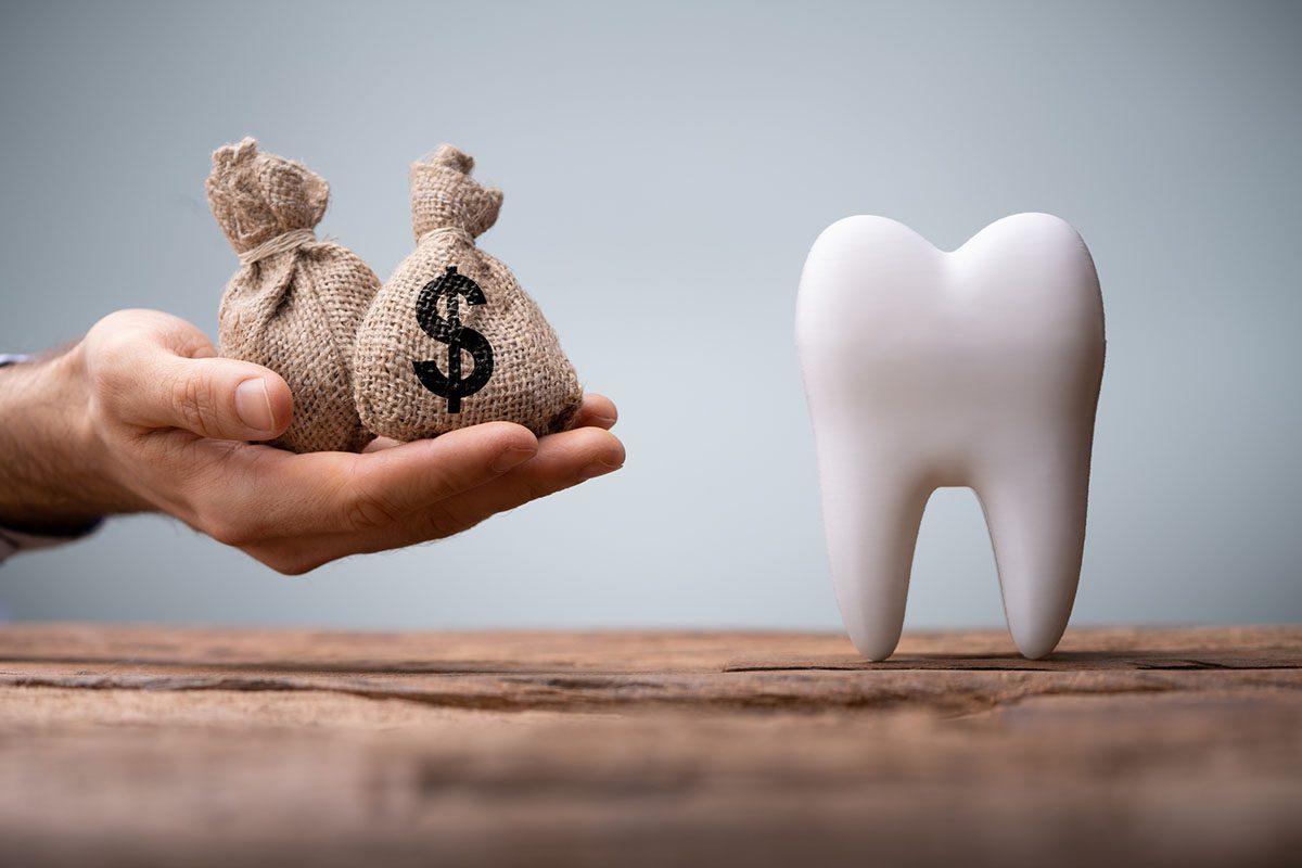 A hand holds two money bags next to a large model of a tooth on a wooden surface with a gray background.