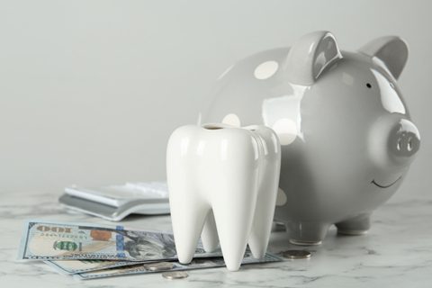 A ceramic tooth and a gray polka dot piggy bank are placed on a marble surface with cash and coins underneath.