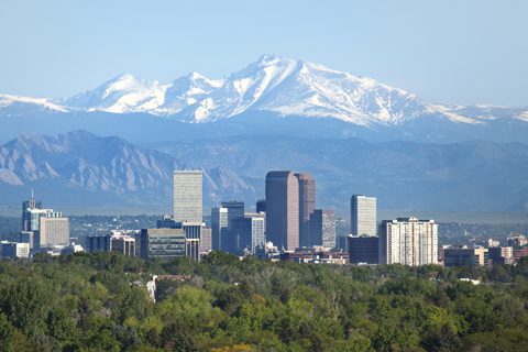 Denver city skyline with tall buildings in the foreground, trees below, and snow-capped mountains in the background under a clear sky.