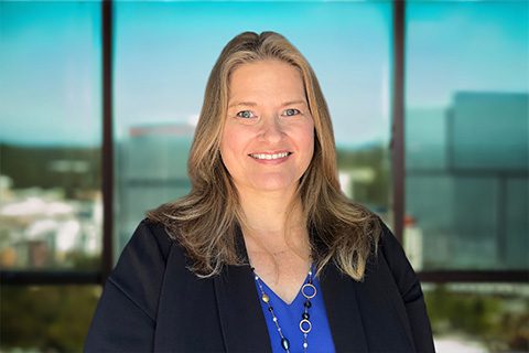 Woman in a black blazer smiles in front of a bright window with a blurred city view.