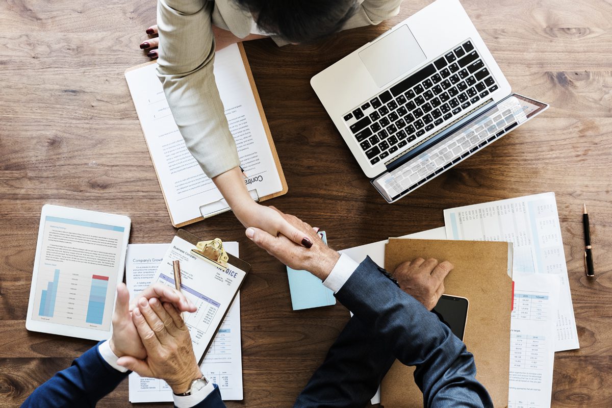 Three people sit at a wooden table with documents and a laptop, as two of them shake hands, suggesting a business agreement or meeting.