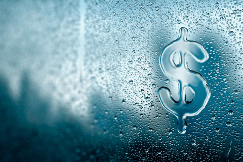 A dollar sign is drawn on a wet, foggy window with water droplets, with a blurred outdoor scene visible in the background.