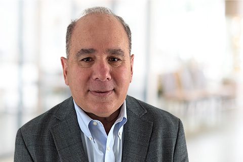 A middle-aged man wearing a gray blazer and light blue shirt, standing indoors with a blurred background.