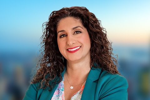 Woman with curly brown hair wearing a teal blazer and floral top, smiling in front of a blurred cityscape background.