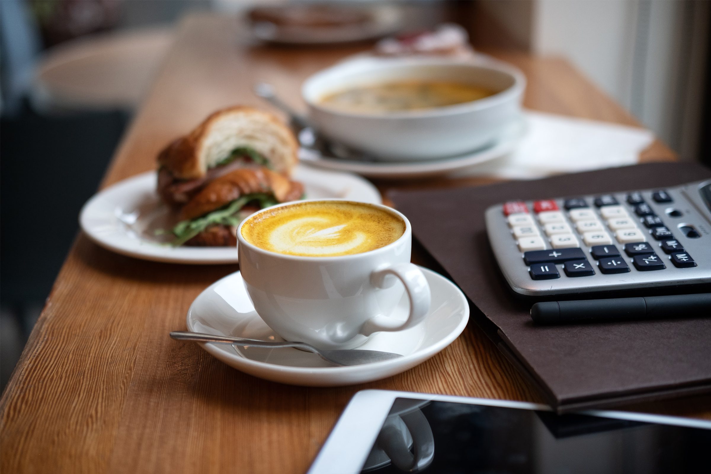 A cup of coffee with latte art sits on a saucer with a spoon, next to a sandwich, a bowl of soup, a calculator, a notebook, and a tablet on a wooden table.