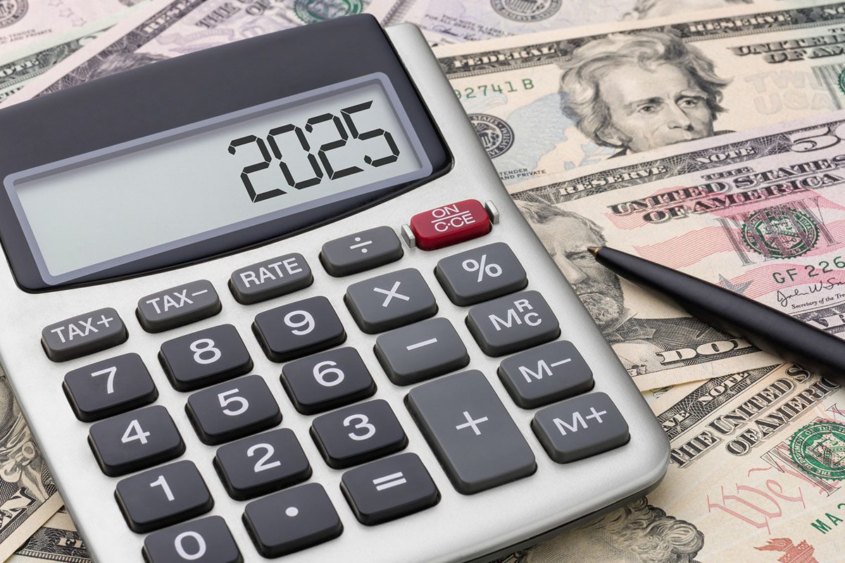 A calculator displaying 2025 sits on top of assorted U.S. dollar bills, next to a black pen.