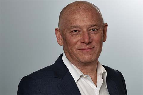 Bald man wearing a dark suit jacket and white collared shirt, smiling slightly, posed in front of a plain light grey background.