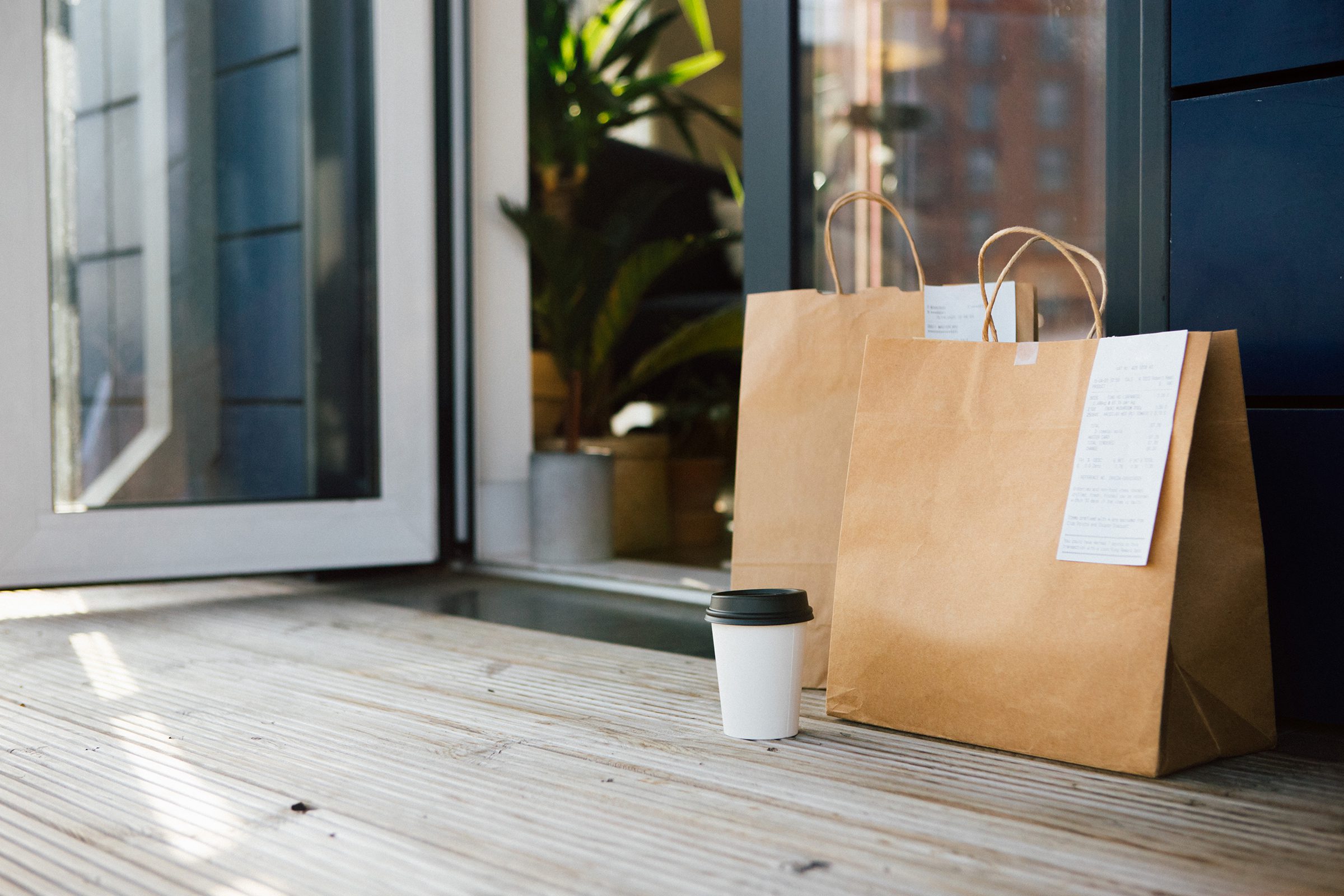 Two brown paper bags with receipts attached and a disposable coffee cup are placed on a wooden porch outside an open door.