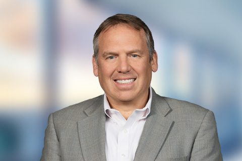 A middle-aged man in a gray suit jacket and white shirt smiles at the camera against a blurred indoor background.