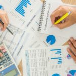 Two people review financial charts, graphs, and reports on paper with a calculator and a notebook on a desk, analyzing data and taking notes.