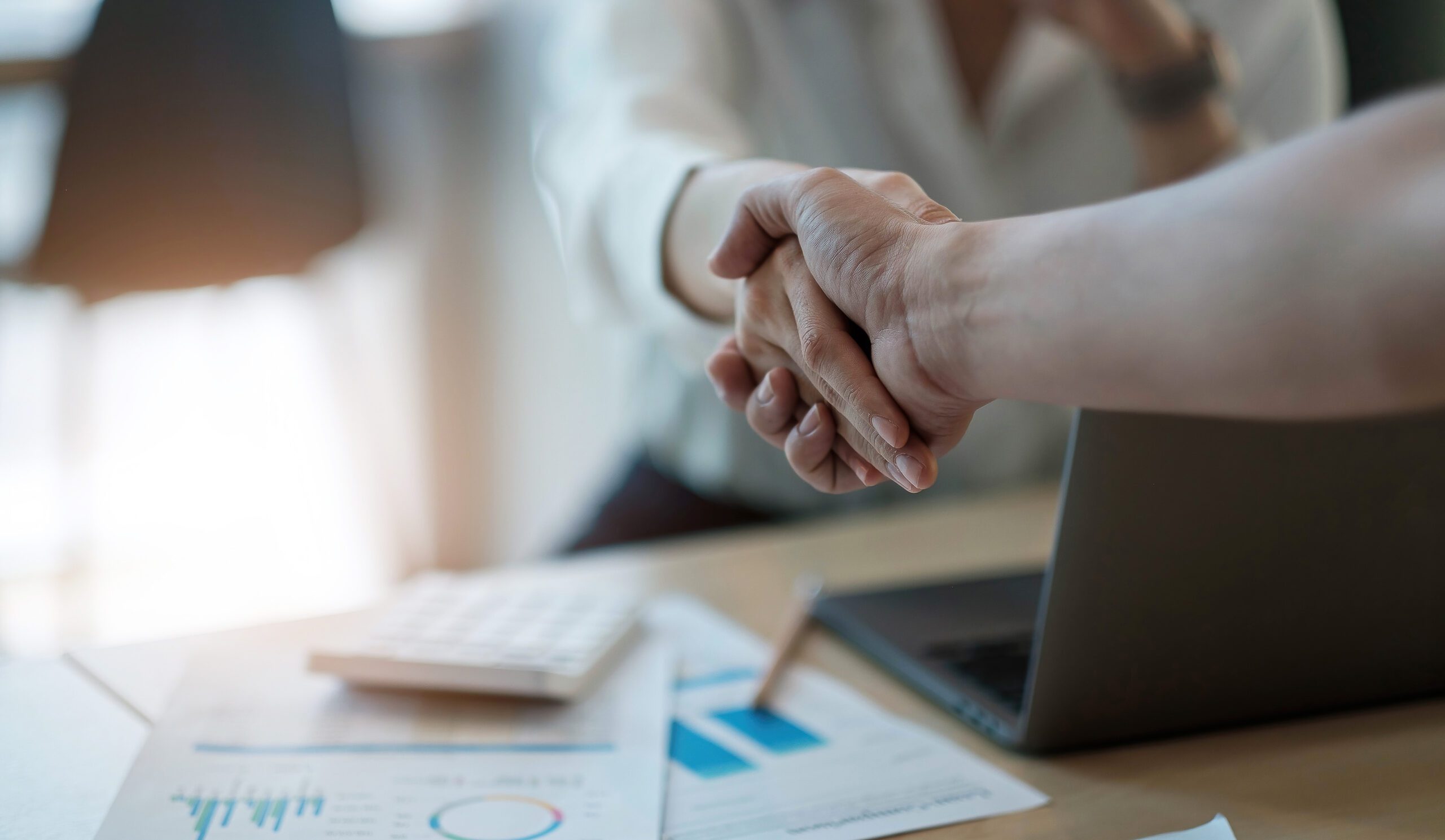 Two people shake hands over a desk with documents, a laptop, and a calculator, suggesting a business agreement or professional meeting.