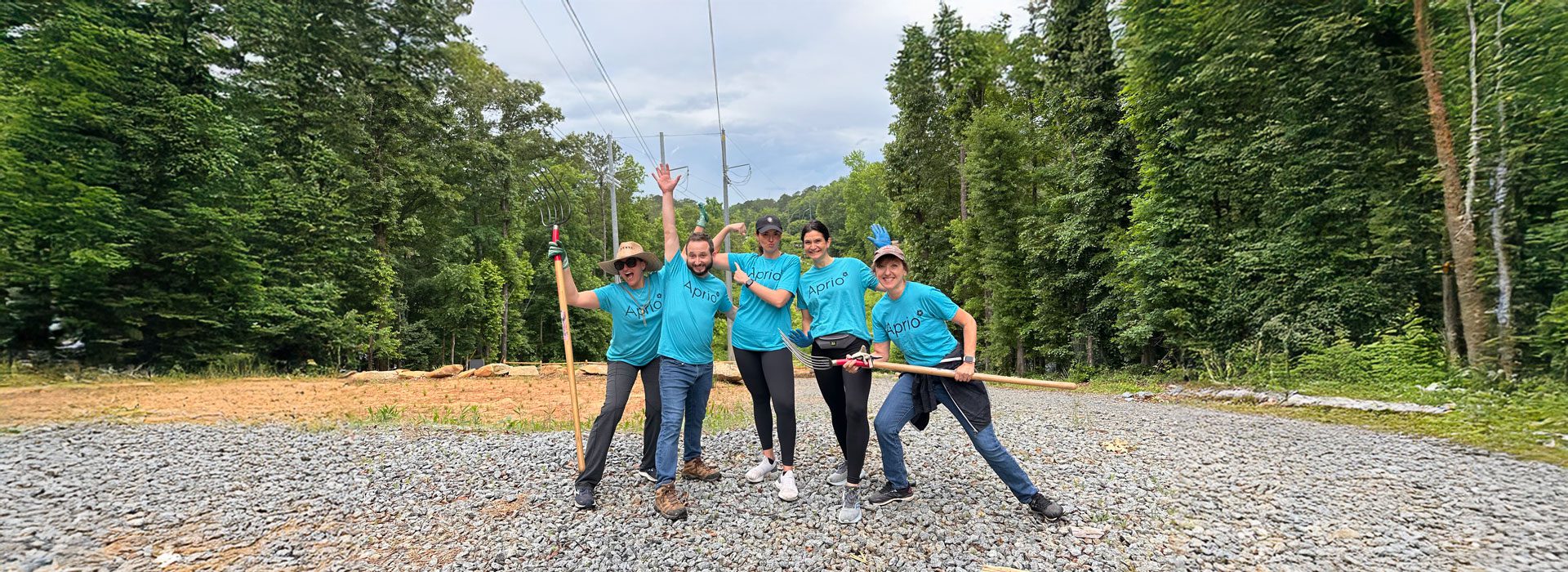 Five people wearing turquoise shirts pose with gardening tools on a gravel path in a forested area under power lines.
