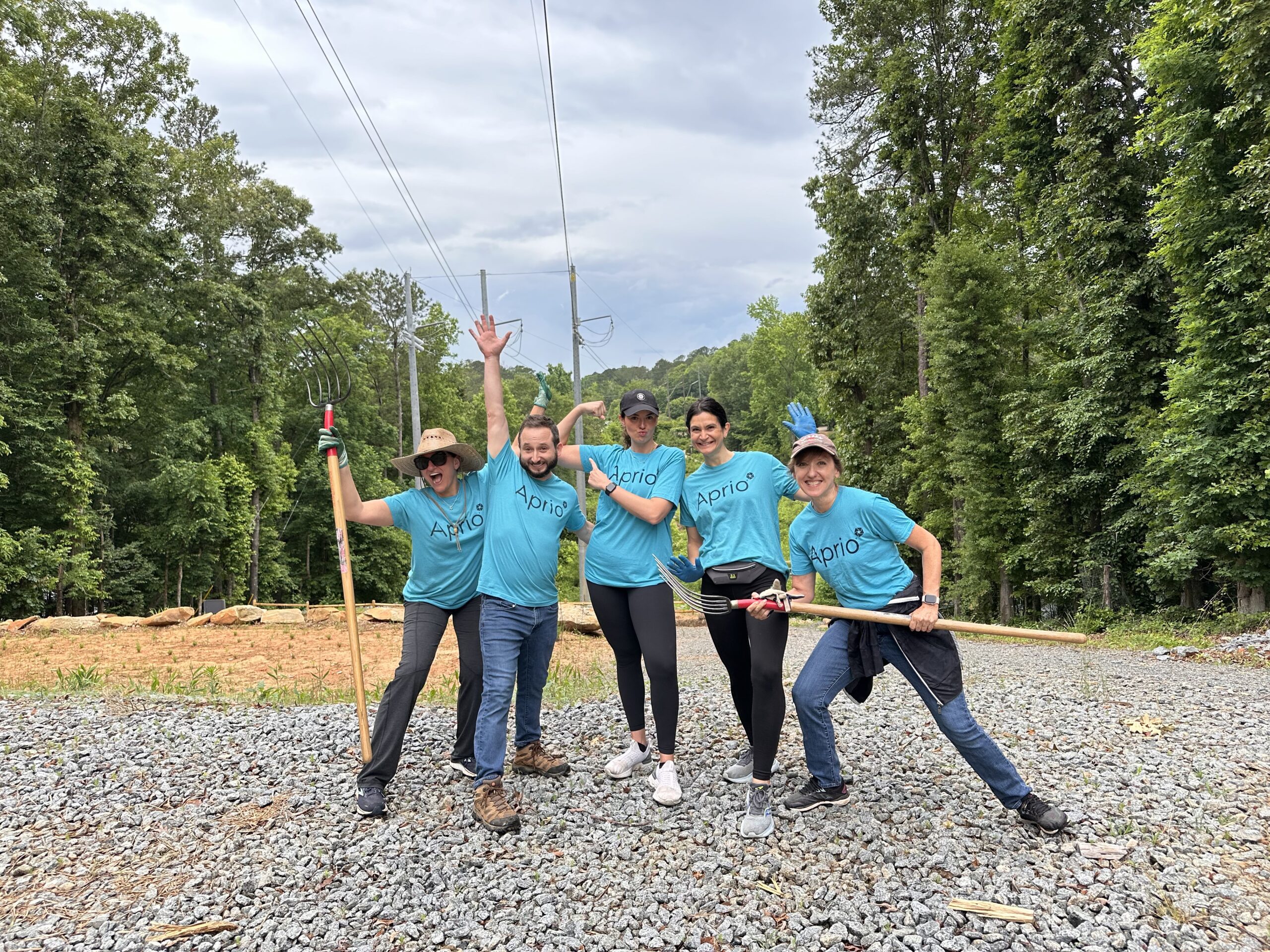 Five people in matching blue shirts stand on gravel in a wooded area, smiling and holding gardening tools, appearing to participate in an outdoor volunteer activity.