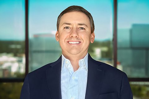 Man in a blazer smiles in front of a bright window with a blurred city view.