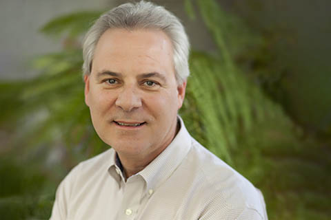 A middle-aged man with short gray hair wearing a light-colored collared shirt, smiling slightly, with green foliage in the blurred background.