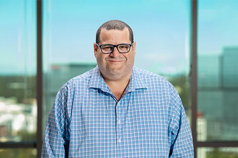 Man in glasses and a blue checkered shirt smiles in front of a bright window with a blurred city view.
