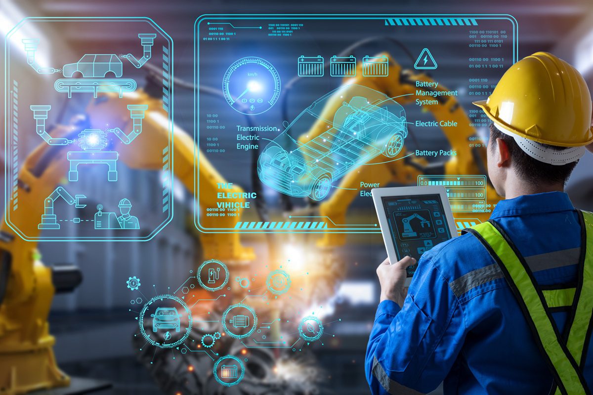 Engineer in hard hat uses a tablet to monitor robotic arms with digital diagrams of an electric vehicle and battery management system displayed as holograms.