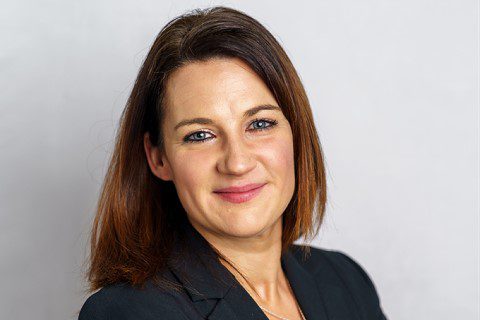 Woman with straight brown hair wearing a black blazer, smiling at the camera against a plain light background.