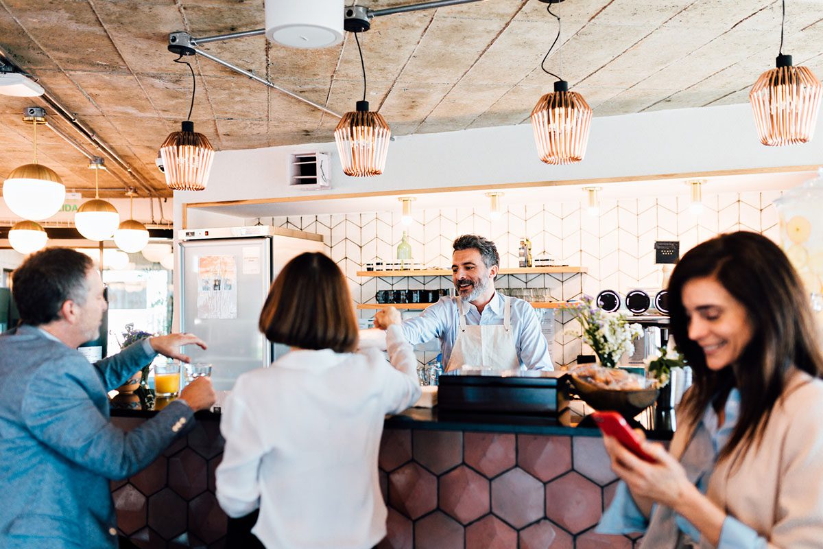 Barista stands behind a café counter smiling and serving drinks to customers; a woman in the foreground uses her phone.