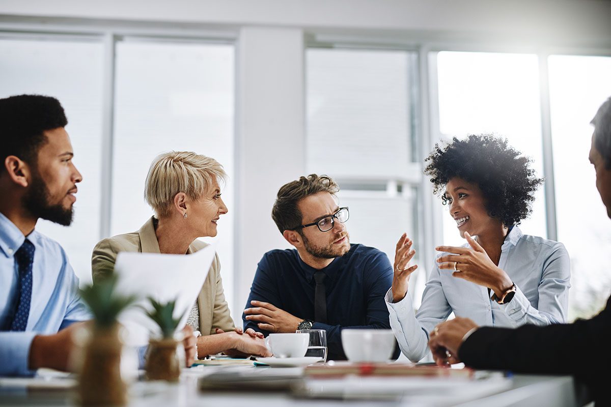 Five people sit around a conference table engaged in discussion; one woman gestures while speaking, and others listen attentively.