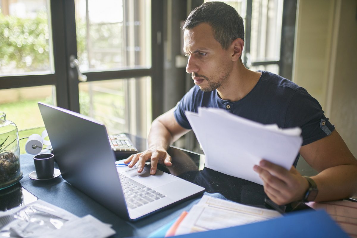 A man sits at a desk using a laptop and holding documents, with papers, a cup, and a calculator visible on the desk in a well-lit room.