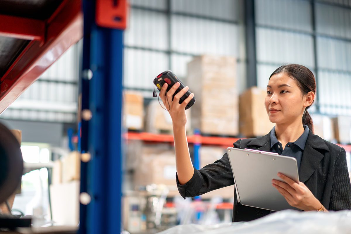A woman in business attire scans inventory with a handheld device while holding a clipboard in a warehouse setting.