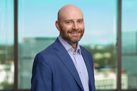 Bearded man in a blue blazer smiles in front of a bright window with a blurred city view.