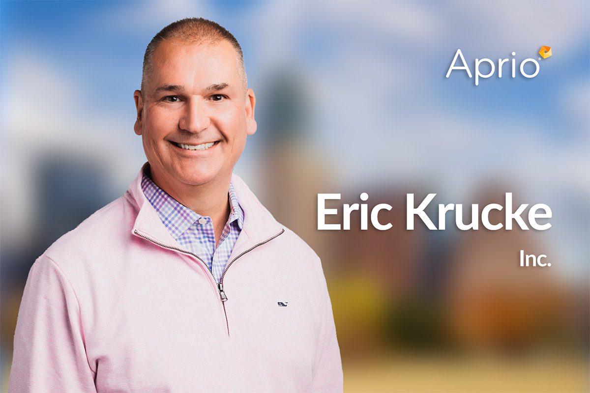 A man wearing a light pink sweater smiles at the camera, with the text Eric Krucke Inc. and the Aprio logo displayed over a blurred cityscape background.