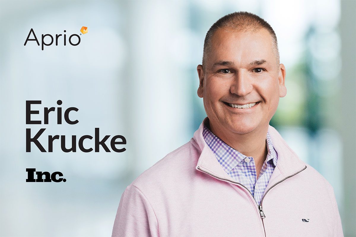 A man in a light purple sweater smiles at the camera. The text reads “Aprio,” “Eric Krucke,” and “Inc.” on a blurred background.