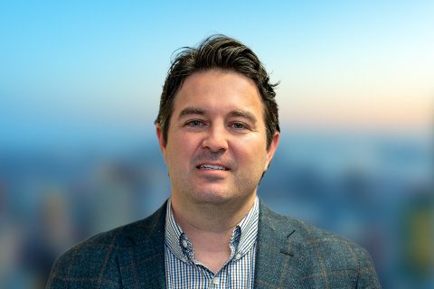 Dark haired man in a blazer appears on a blurred bright background