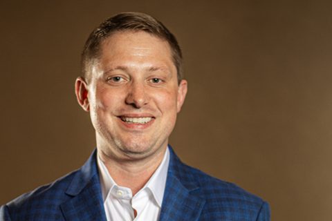 A man wearing a blue plaid jacket and white shirt smiles in front of a plain brown background.