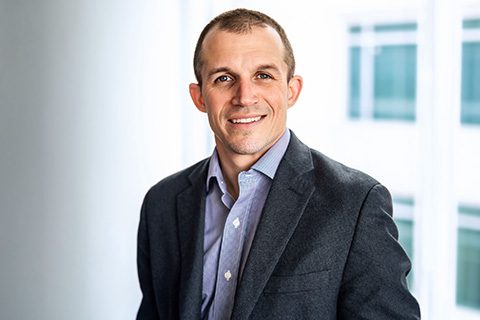 Man in a gray suit jacket and light blue shirt stands indoors, smiling, with a blurred windowed background.
