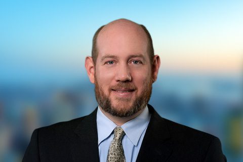 Bearded man in a blazer smiles in front of a bright blurred background