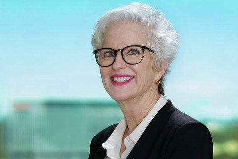 An older woman with short white hair, glasses, and a black blazer smiles while standing in front of a blurred outdoor background.