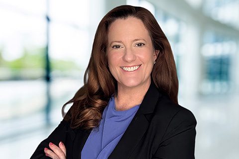 Dark haired woman in a blazer smiles in front of a bright blurred lobby background