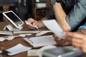 Two people review paper receipts and documents at a table next to a tablet device, with various papers scattered around.