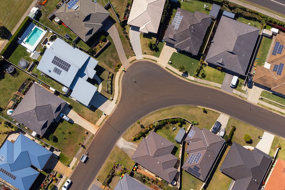 Aerial view of a suburban neighborhood showing several houses with driveways, lawns, solar panels, and a curved road running through the center.